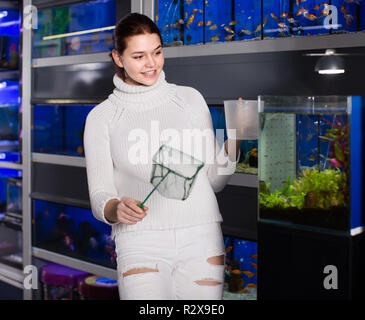 happy american girl holding aquarian net and water container next to aquarium with colorful fish in aquarium shop Stock Photo