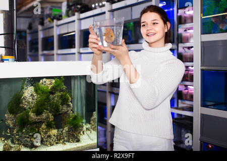 happy european girl holding plastic container with big colorful fish breed Discus in aquarium shop Stock Photo