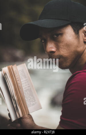 Adventurer guy relaxing and reading a book in the woods next to a river Stock Photo