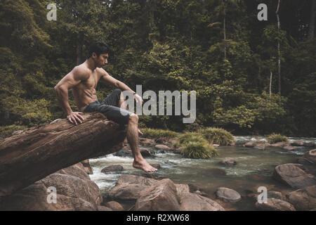 Muscular man sitting on a tree trunk while enjoying the nature around him Stock Photo