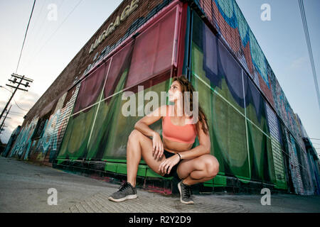 Female athlete kneeling on sidewalk in front of boarded up building covered in graffiti. Stock Photo