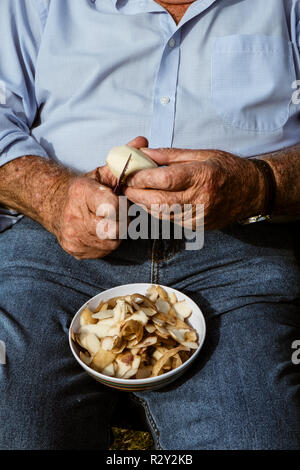 A man seated using a sharp knife to peel potatoes. Stock Photo