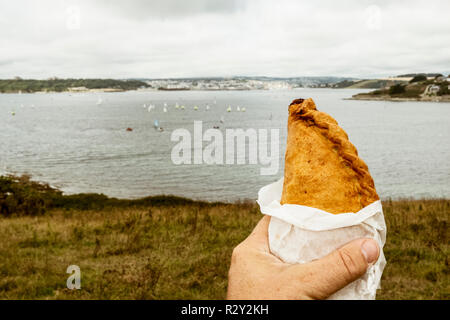 A hand holding a Cornish pasty baked pastry with a crimped edge, seated overlooking a sea estuary. Stock Photo