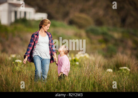 Little girl and her pregnant mother chat together as they walk hand in hand through a grassy meadow. Stock Photo