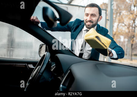 Smiling businessman dressed in a suit wiping windshield of his car with yellow microfiber on a self service car wash