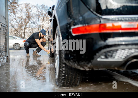 Professional washer in black uniform and cap wiping with sponge car wheel of a luxury car during the washing process outdoors Stock Photo