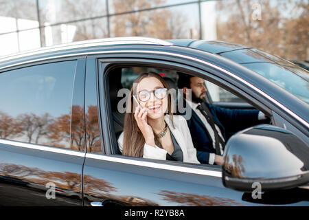Elegant businesswoman talking with phone driving luxury car with businessman in the city, view from the outside through the window Stock Photo