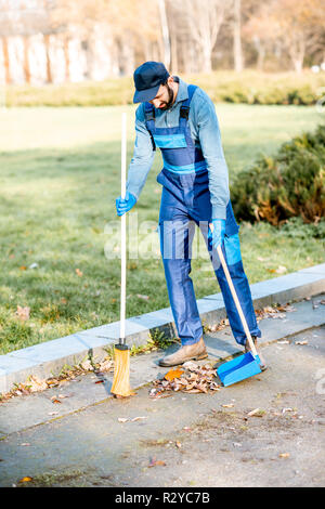 Professional sweeper in uniform sweeping leaves with broom and scoop on the street Stock Photo