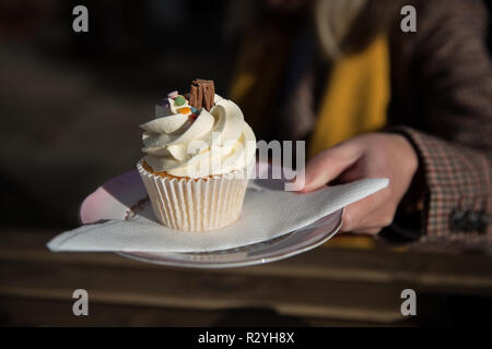 A close up of a girls hand holding up a delicious vanilla cupcake or muffin with swirled frosting on a plate and napkin with copy space Stock Photo