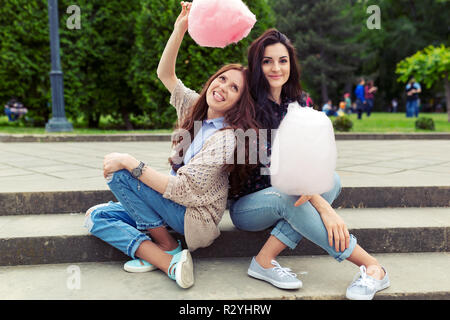 Young, cheerful girls having fun with cotton candy outdoors on urban background. Two sisters making funny faces and mustache while playing and eating  Stock Photo