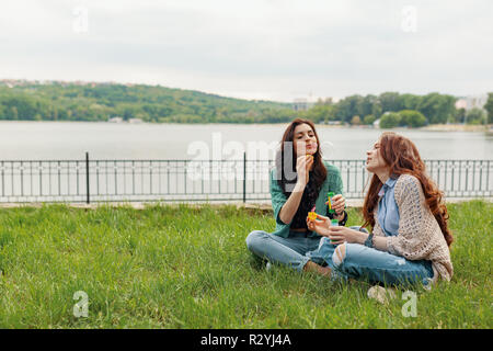 Two sisters having fun while making bubbles and sitting on the grass near the lake with a beautiful landscape in the background Stock Photo