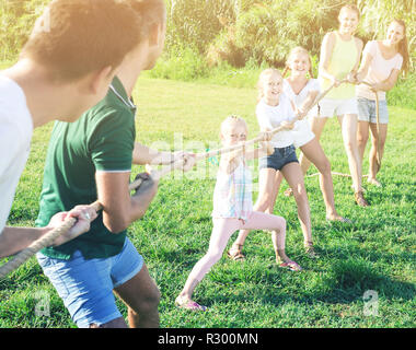 Ordinary kids with moms and dads playing tug of war during joint outdoors games on sunny day Stock Photo