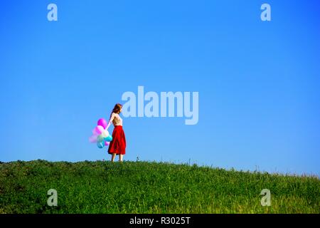 Happy girl in the meadows tuscan with colorful balloons, against the blue sky and green meadow. Tuscany, Italy Stock Photo