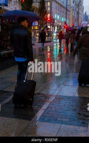 A man pulling  a trolley suitcase in Gran Via, Madrid during a wet and dark late afternoon on Sunday. Stock Photo