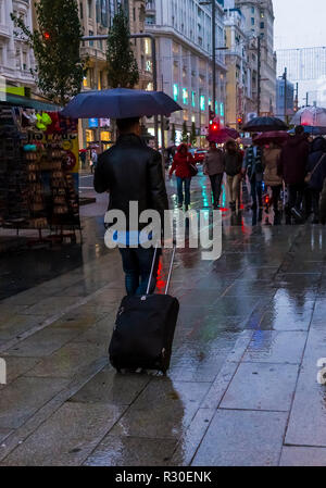 A man pulling  a trolley suitcase in Gran Via, Madrid during a wet and dark late afternoon on Sunday. Stock Photo