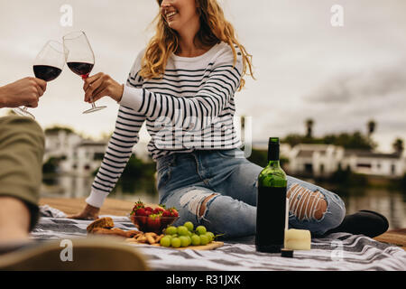 Couple on a date sitting beside a lake eating snacks and drinking wine. Young woman sitting with her boyfriend drinking wine on a date. Stock Photo