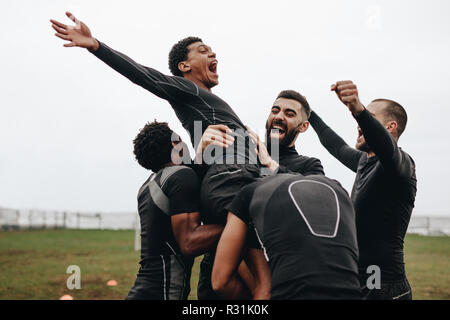 Group of soccer players celebrating a goal standing together on field. Teammates celebrating a goal by lifting a player and shouting in joy. Stock Photo