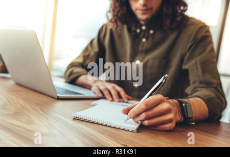 Entrepreneur making notes sitting at his work desk in office. businessman sitting with a laptop on table writing notes in a book. Stock Photo