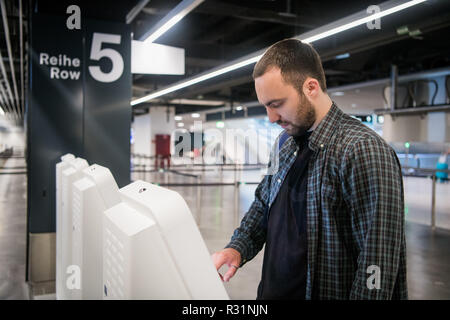 Young man with backpack touching interactive display using self service machine, doing self-check-in for flight or buying airplane tickets at automatic device in modern airport terminal building Stock Photo