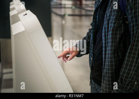 Young man with backpack touching interactive display using self service machine, doing self-check-in for flight or buying airplane tickets at automatic device in modern airport terminal building Stock Photo