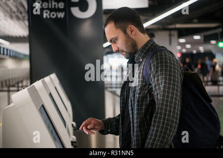 Young man with backpack touching interactive display using self service machine, doing self-check-in for flight or buying airplane tickets at automatic device in modern airport terminal building. Stock Photo