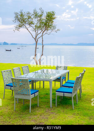 Dining table set with Seascape background. Modern table and chairs with blue pad, dining table set on green lawn with mangrove tree, boat, island and  Stock Photo