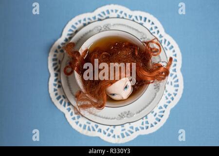 Surreal image of red-headed doll in a cup of tea. Stock Photo
