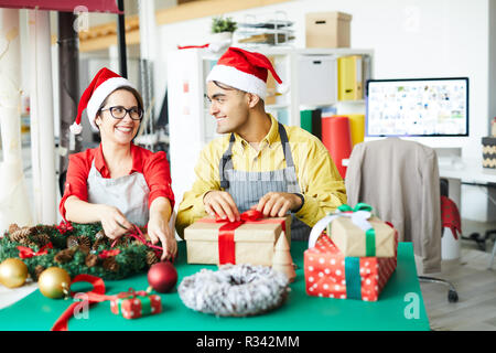 Two young colleagues in Santa caps tying red ribbons on wreath and giftbox while looking at one another Stock Photo