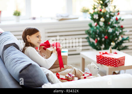 Little girl in casualwear trying to guess what is in giftbox while slightly shaking it Stock Photo