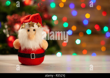 The Christmas toy Santa Claus with three gold stars is in a cap on a light table against the background of the shining multi-colored garland Stock Photo