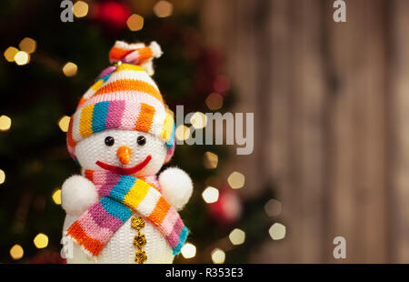 Snowman smiling in multi-colored hat and scarf Christmas toy on the background of a Christmas tree with a garland and a wooden wall Stock Photo