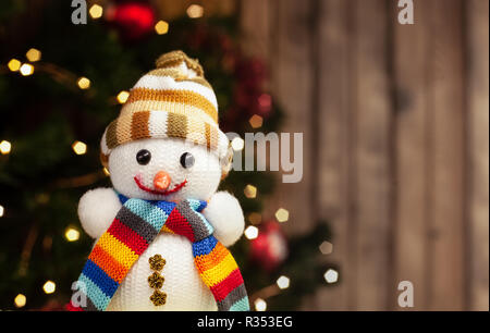 Snowman smiling in multi-colored hat and scarf Christmas toy on the background of a Christmas tree with a garland and a wooden wall Stock Photo