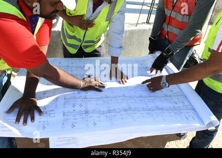 Men with Blueprints in hand are working in the construction Industry and wearing hard hat and safety vest, they stay together under the blue sky and s Stock Photo