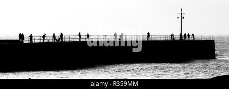 Black and white panoramic image of the harbour wall at Portreath with several people spread along the length looking at the seas beyond. Stock Photo