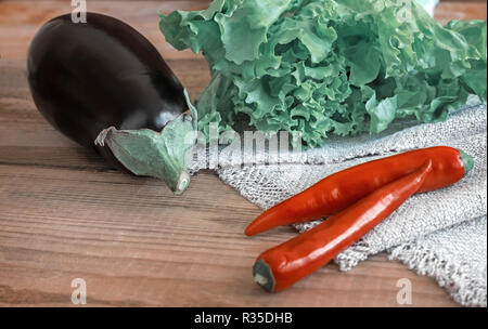 On a wooden table are vegetables: eggplant, chili and green salad in a small red container. Stock Photo