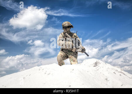 Man in military camouflage uniform with service rifle replica, standing on top of sand dune with cloudy sky on background, imitating U.S. army special Stock Photo