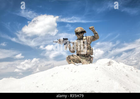 Airsoft military games player in camouflage uniform and helmet armed assault rifle replica, sitting on knee on top of sand dune with sky on background Stock Photo