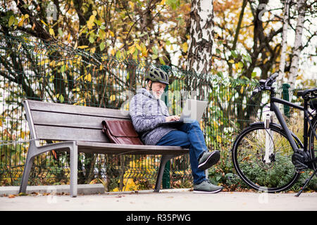 A senior man with electrobike sitting on a bench outdoors in town, using laptop. Stock Photo