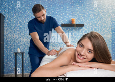 Front view of woman lying on couch, looking at camera and smiling during creole massage in spa salon. Masseur in uniform massaging legs of woman with bamboo sticks. Concept of enjoying procedure. Stock Photo