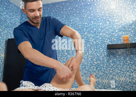 Smiling masseur doing anticellulite massage to woman lying on couch in spa cosmetology salon. Doctor keeping apparatus in strong hands and massaging hips of female client. Concept of beauty. Stock Photo