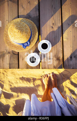 The girl's legs near the hat and two cups of coffee stand on the pier. Stock Photo