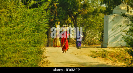 RAJASTHAN INDIA DUSTY ROAD AND THREE WOMEN WITH COLOURFUL SAREES AND LOADS ON THEIR HEADS Stock Photo