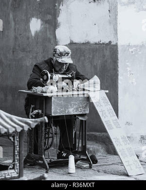 Black and white photo of elderly chinese man earning a living using a sewing machine for clothing alterations in a street in Dali China. Stock Photo