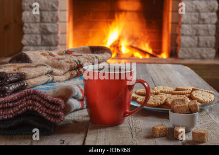 Orange mug for tea or coffee with cookies ans sugar, wool things against cozy fireplace background; winter vacations,horizontal. Stock Photo