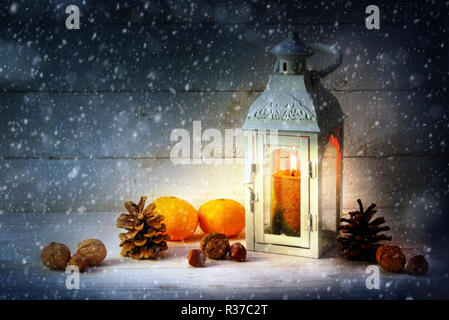christmas decoration with a candle light lantern, tangerines, cones and nuts in the snow on rustic wood at night, copy space, selected focus Stock Photo