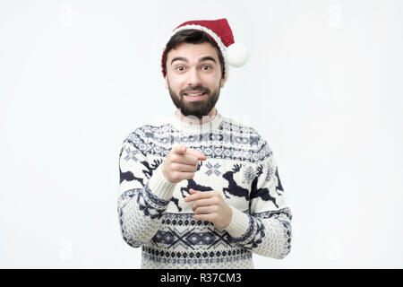 Young hispanic man in red christmas cap pointing at camera laughing. Stock Photo