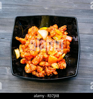 Chinese cuisine dish - top view of black bowl with Fried chicken pieces with pineapple and bell pepper and onion (Sweet and Sour Chicken) on dark wood Stock Photo