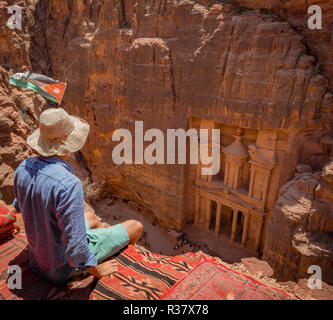 Tourist with sun hat sitting on carpet, looking from above into the gorge Siq, Pharaoh's treasure house carved into rock, facade Stock Photo