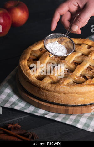 Home baking concept with a woman hands pouring powdered sugar from a sieve to a classic apple pie, on a kitchen table. Traditional sweet food. Stock Photo