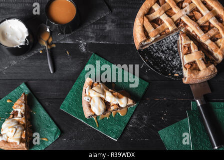 Above view of a rustic table with a sliced apple pie, and two pieces on napkins with cream and caramel sauce. Homemade cake. Traditional classic pie. Stock Photo